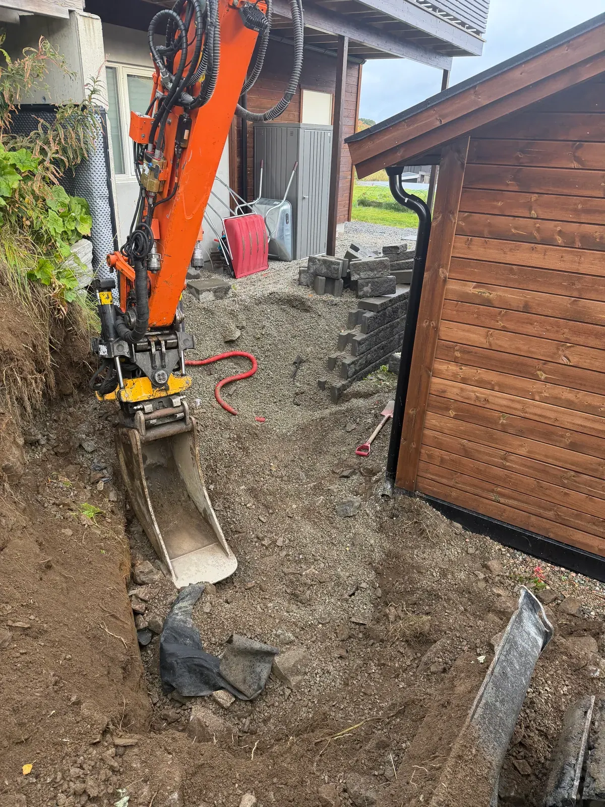 Gravemaskin som graver ut tett inntil en garasjevegg