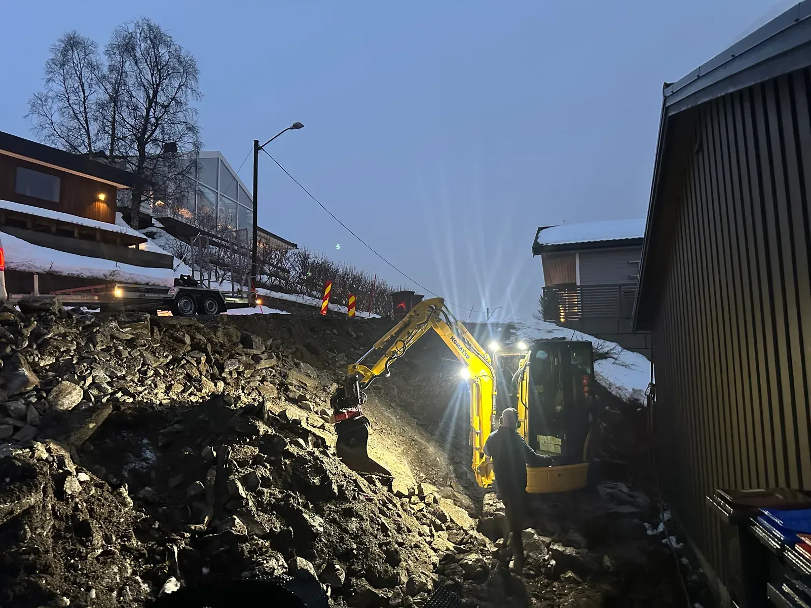 Gravemaskin graver i lyskasterlys mellom to bygg